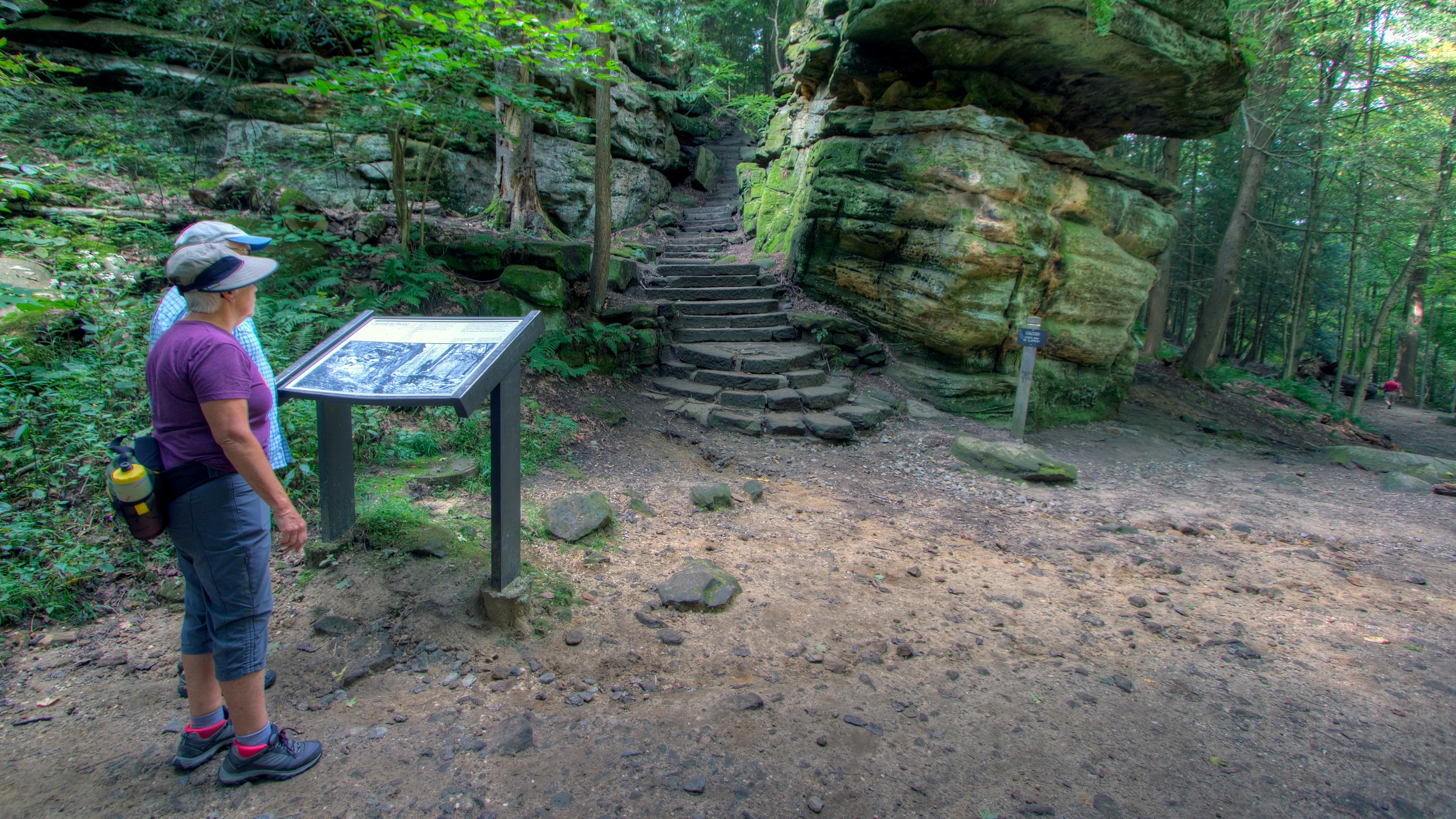 Two people read a sign in front of a rock formation with stairs.