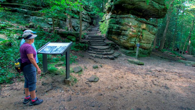 Two people read a sign in front of a rock formation with stairs.