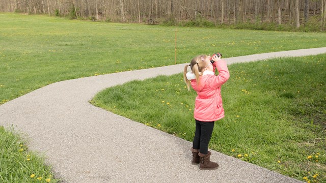 A young girl in a pink coat standing on a path looks off to the right through a pair of binoculars