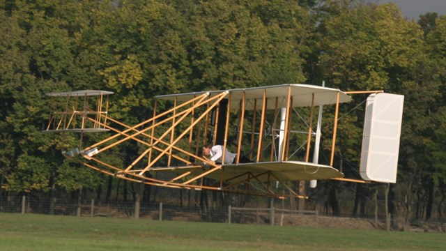 A replica Wright brothers plane flies low to the ground with trees in the background