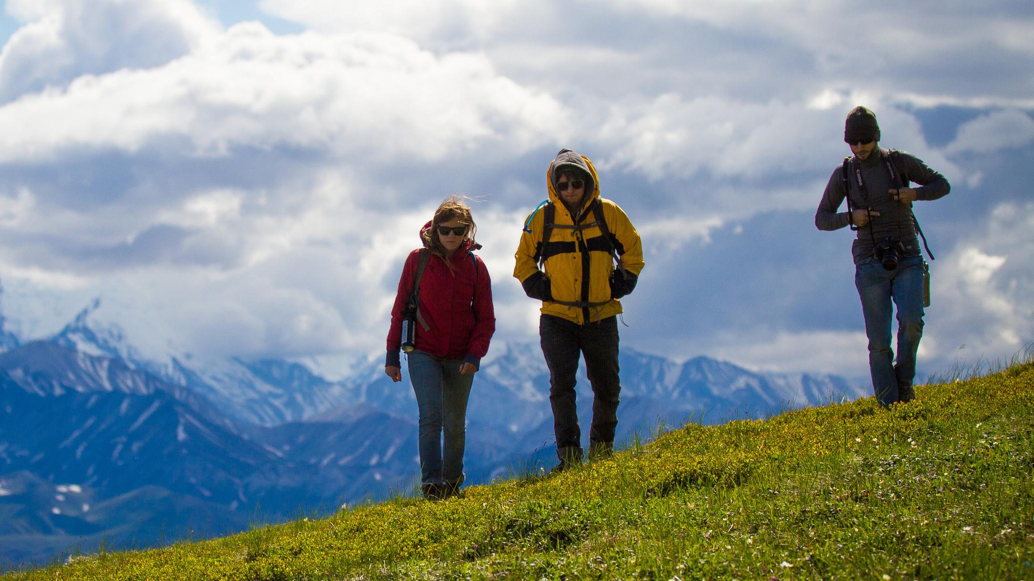 Three hikers on a grassy slope with clouds behind them