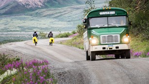 two bicyclists ride on a dirt road in the opposite direction of a bus