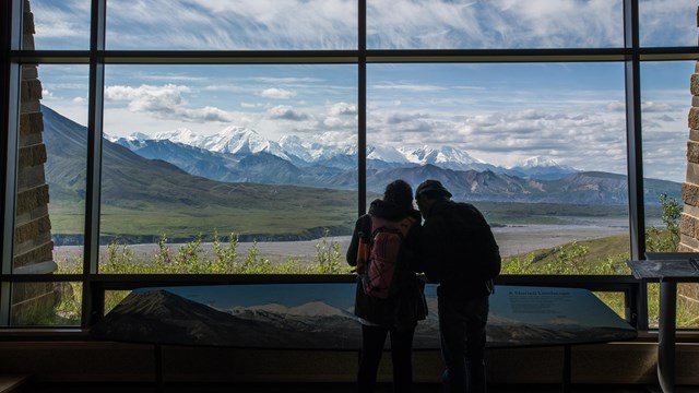 two people standing indoors in front of a window, looking out at mountains