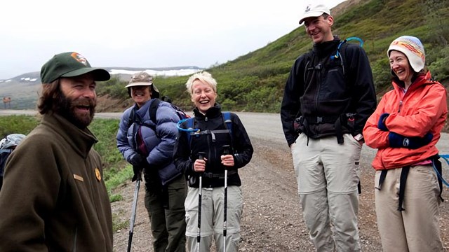 A male ranger and four hikers stand on a dirt road laughing.