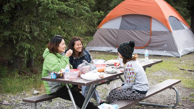 Three women sit at a picnic table, laughing and eating food. Behind them is a large orange tent.