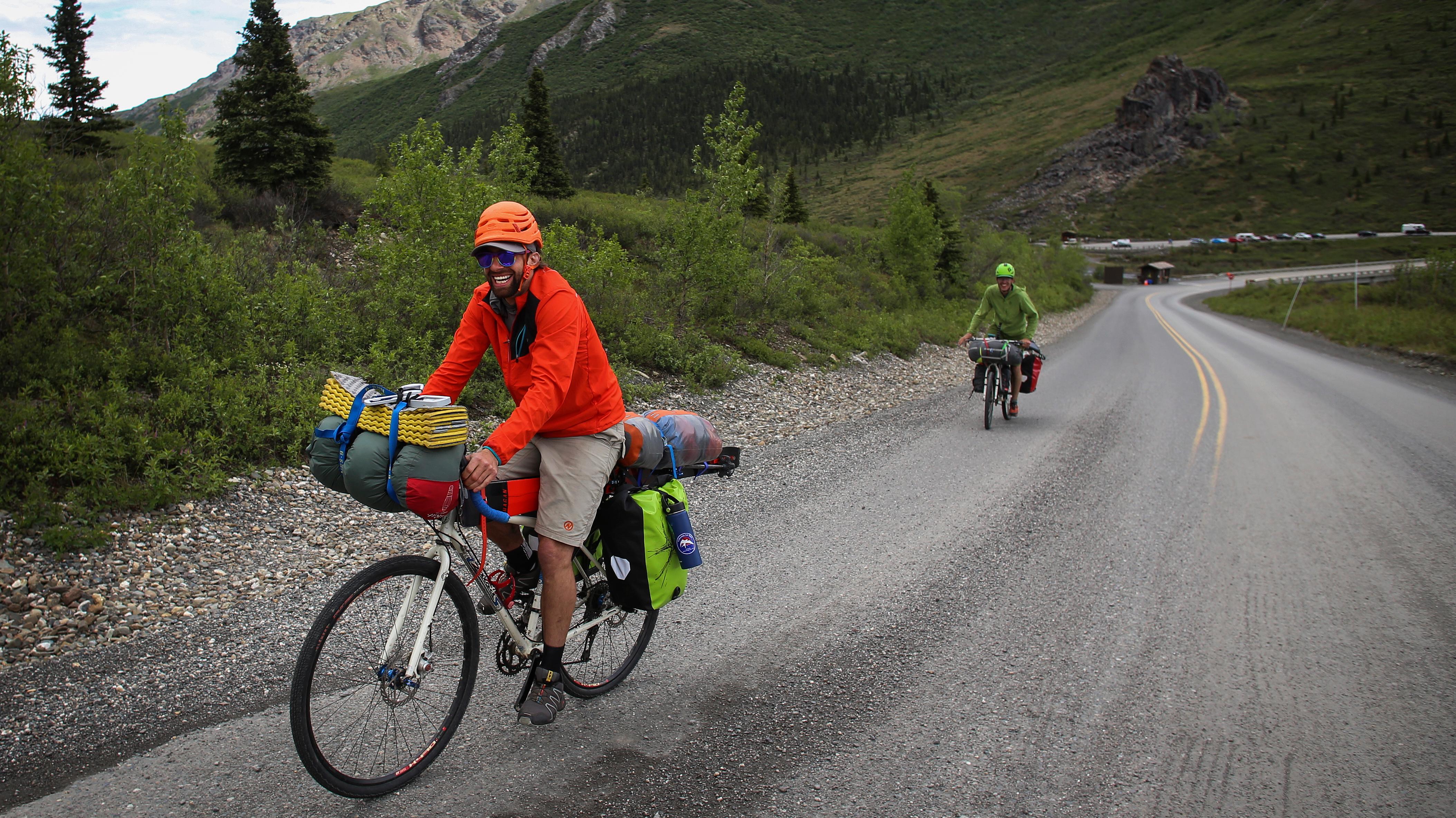 Two smiling cyclists biking uphill with heavily loaded bikes carrying lots of gear.
