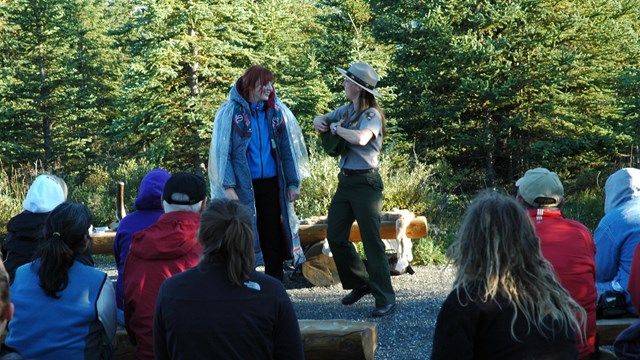 a female ranger speaking to a group of people seated around her in a forest clearing