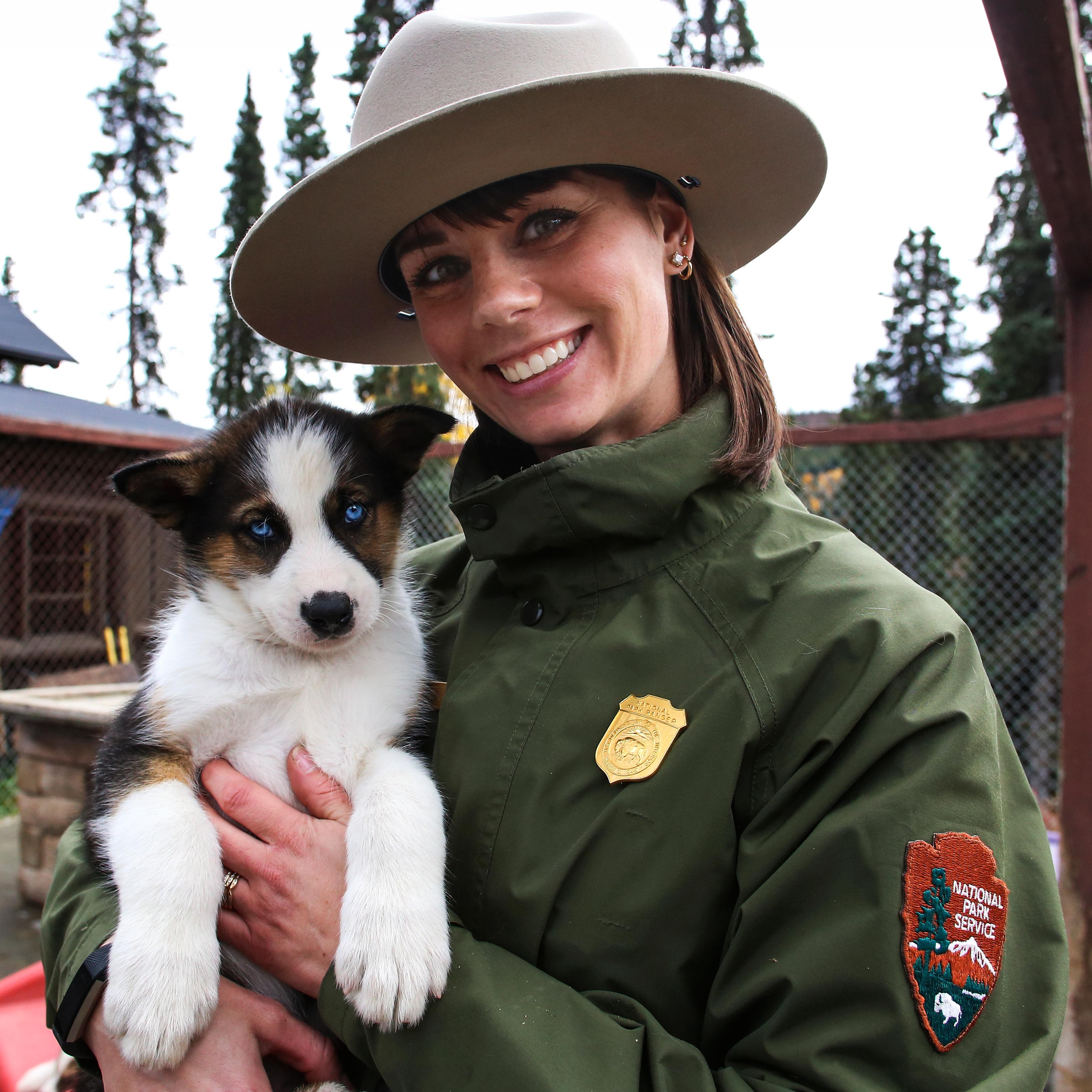 A smiling ranger holds a brown and white puppy in her arms.