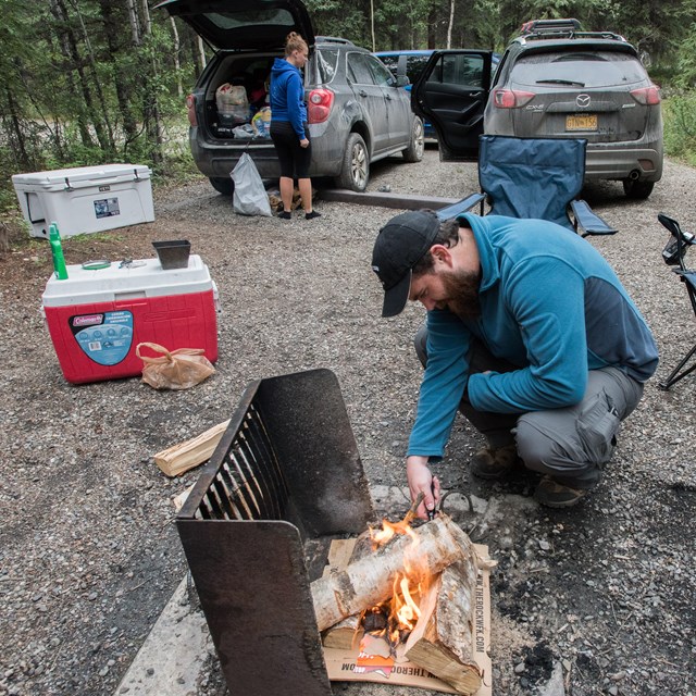 man kindling a fire while a woman unloads camping equipment from a car