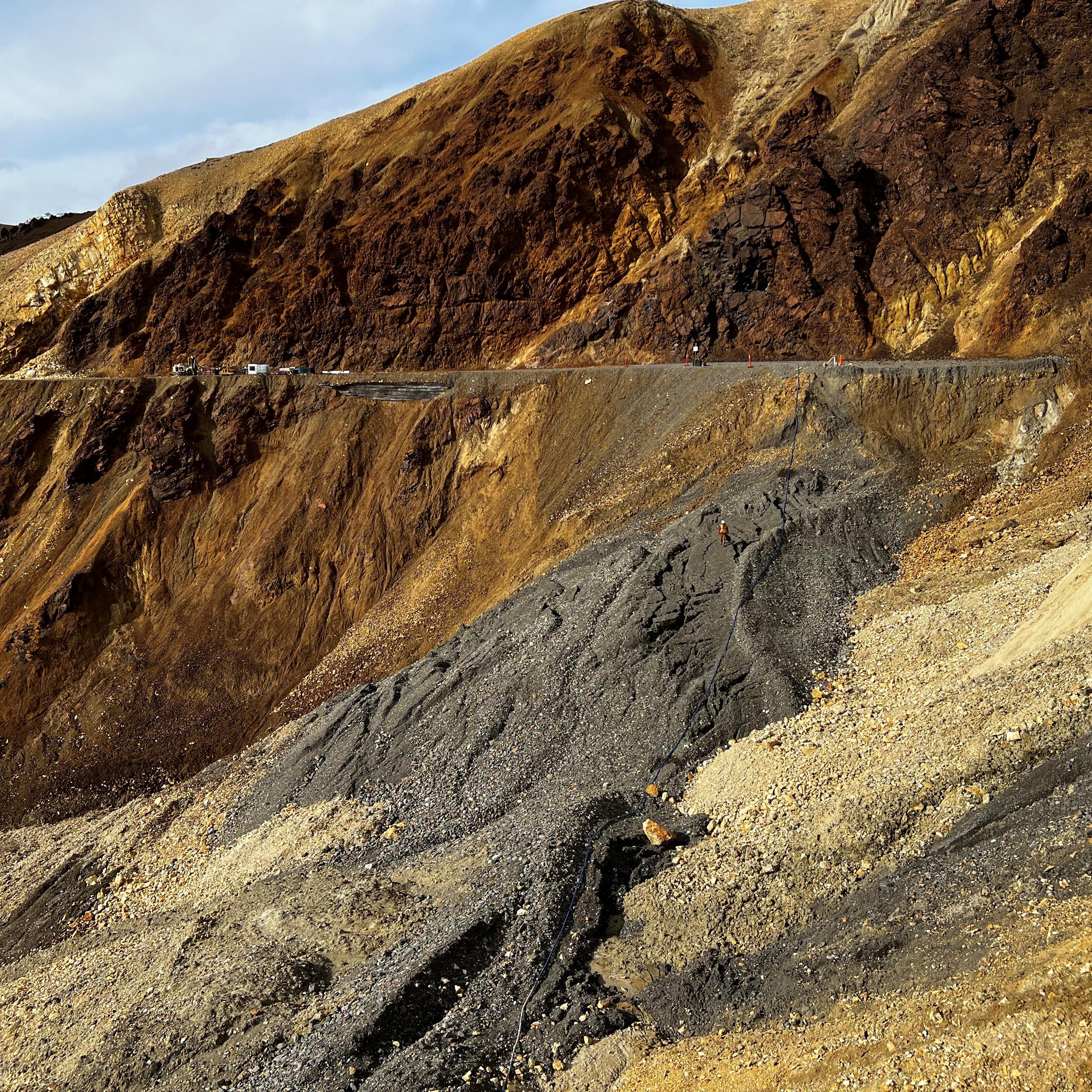 A mountainside of orange and gray rock slumping downhill away from the flat road bed.