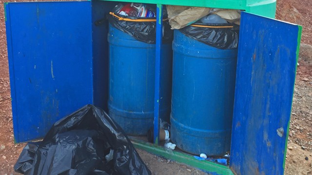 Two trash cans, full, with a black trash bag in front. 