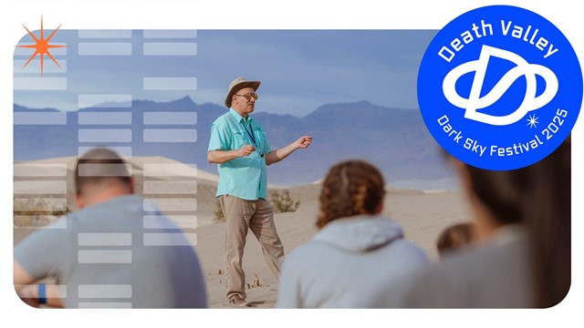 A man standing on sand dunes talks to a crowd with mountains in the background.