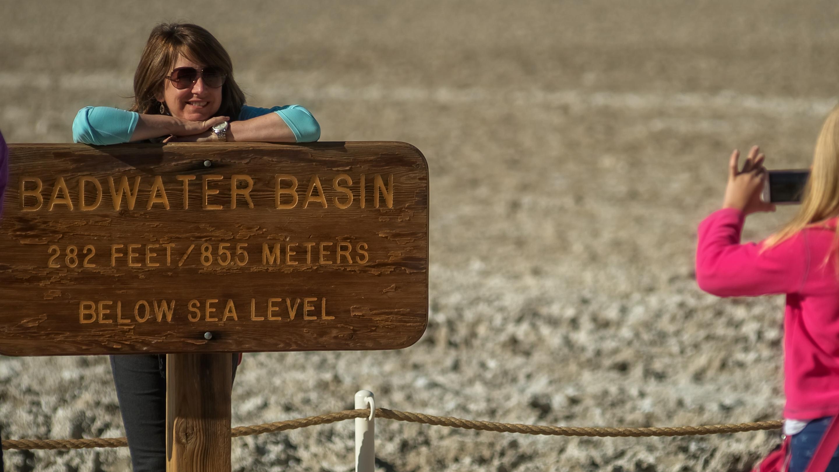 A child takes a picture of a woman at the wooden Badwater sign reading: 282 feet below sea level