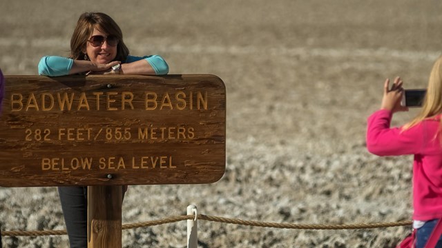 A child takes a picture of a woman at the wooden Badwater sign reading: 282 feet below sea level