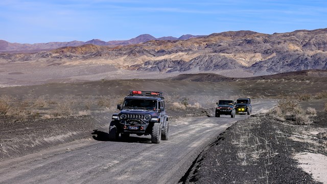 Three jeeps drive down a dirt road in a desert mountain landscape.
