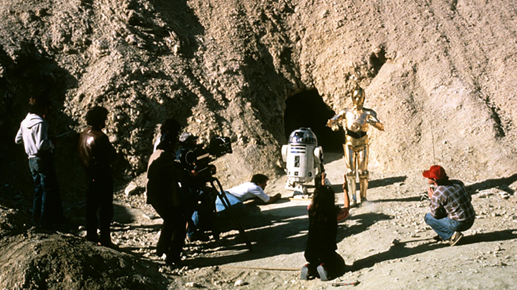 Two robots are surrounded by a few people filming them in a desert landscape. 