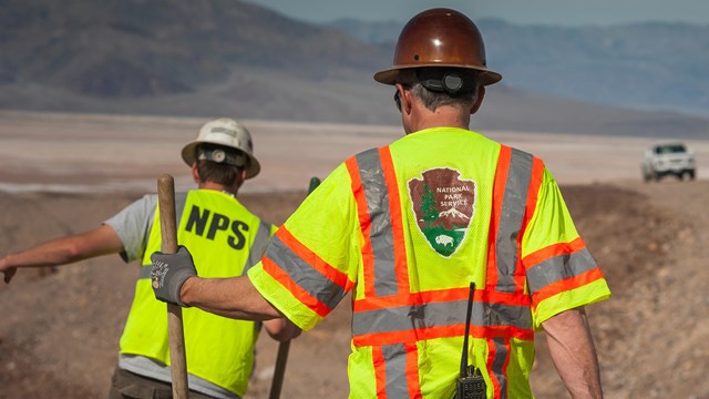  National Park Service workers in bright safety vests tend to a dusty road construction site.
