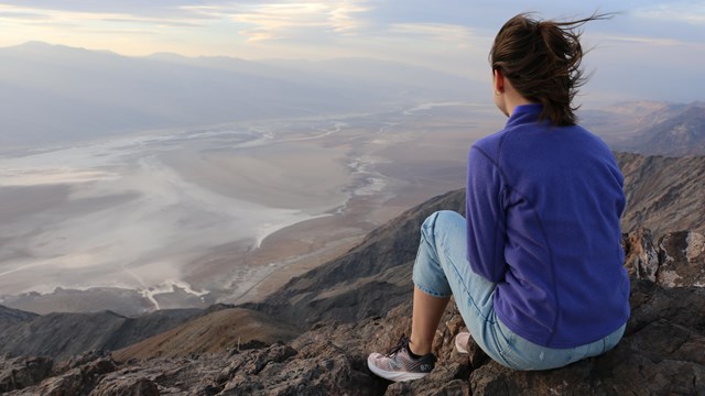 A woman sits on a rock ledge overlooking a valley of white swirling salt with pastel clouds above.