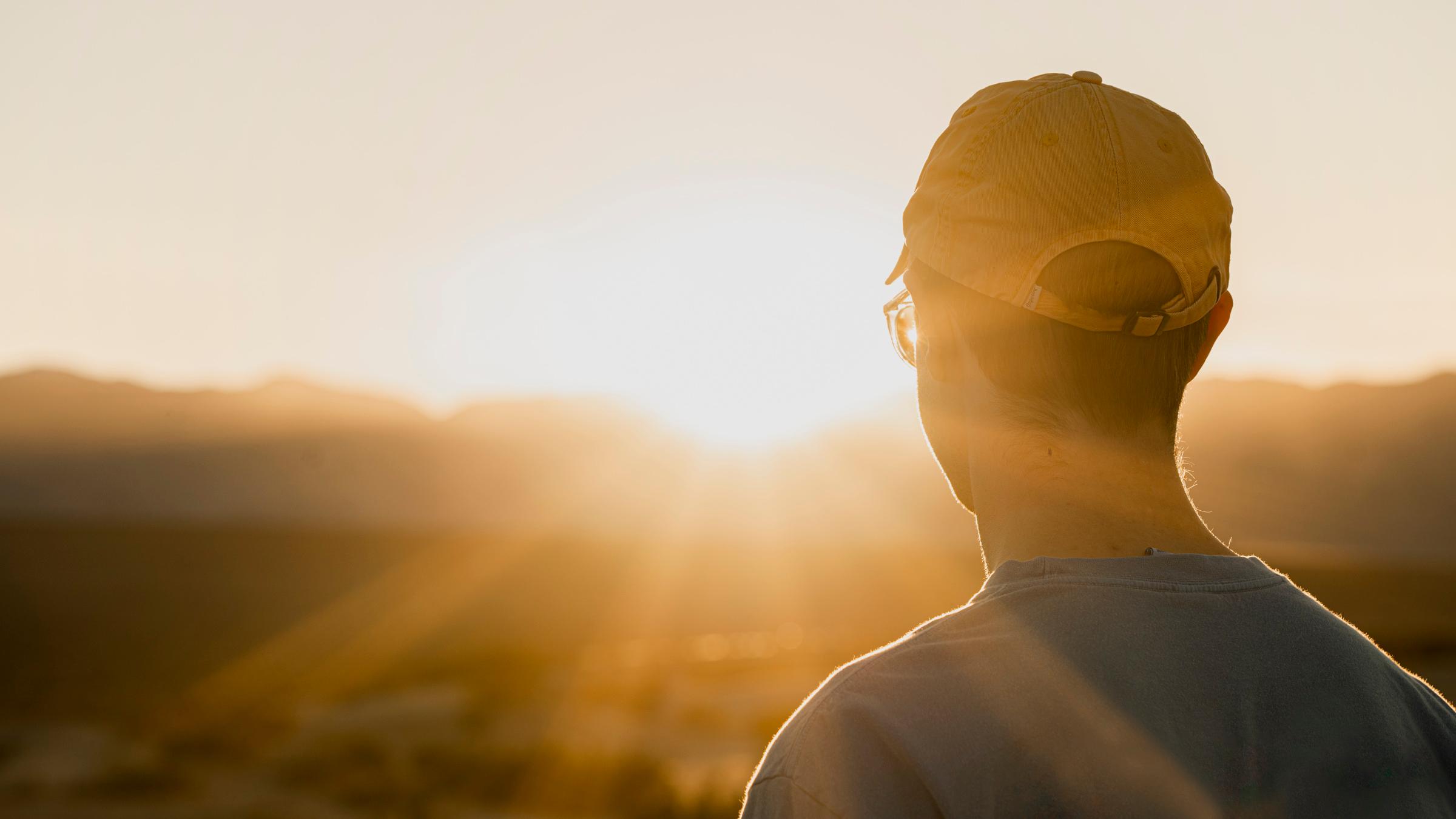 An individual in a t-shirt and ball cap watches the sunset over the distant mountains as light beams