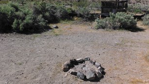 A rock fire ring with ruins of a wooden and metal structure and desert shrubs in the background.