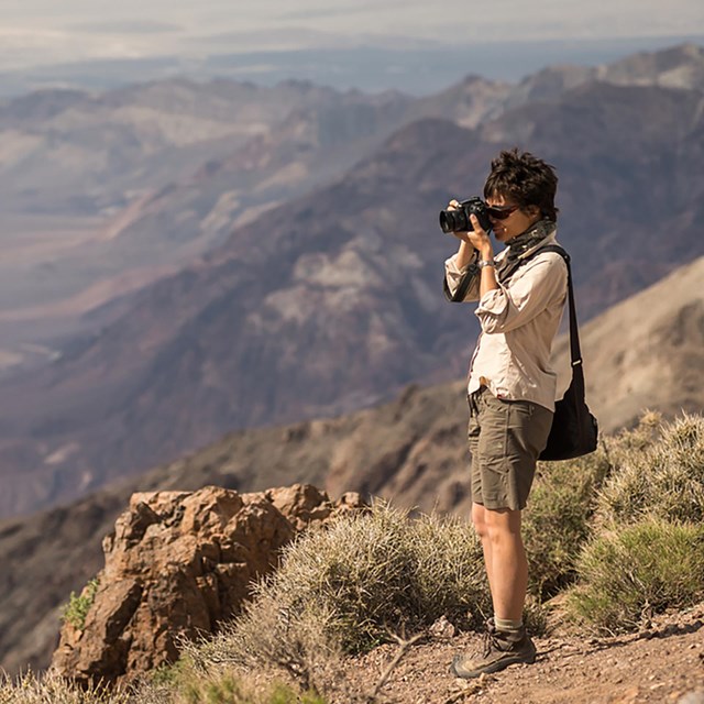 A photographer with camera from the side from a mountain overlook, with rocky slopes.