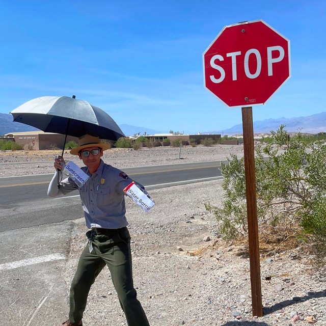 A park ranger stands beside a red STOP sign on a sunny day, holding an umbrella 