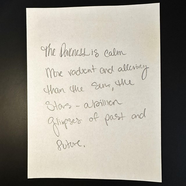 A white piece of paper on a black background. Written in pencil on the paper is a poem.