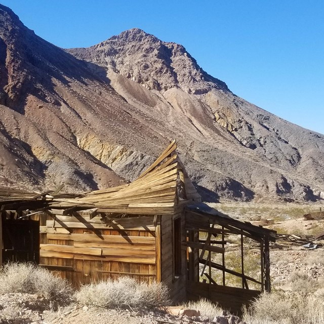The remains of a weathered wooden building with a sagging roof stands before the remains.