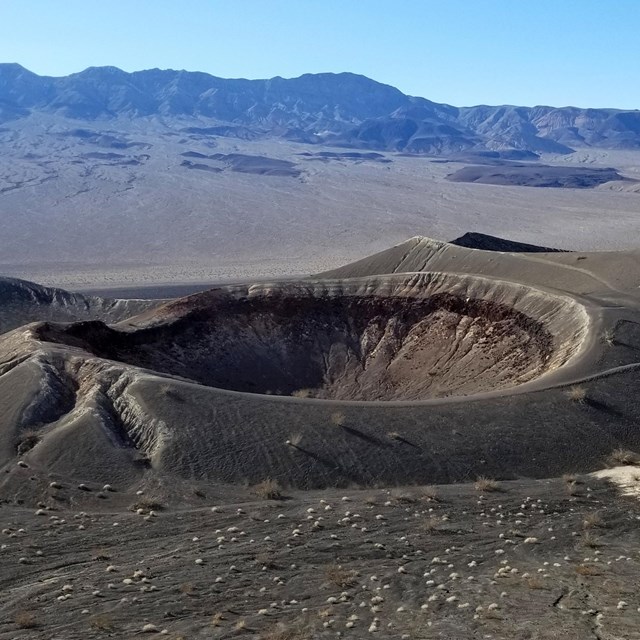 A path leads around the rim of a crater in a landscape of grey cinders with mountains.