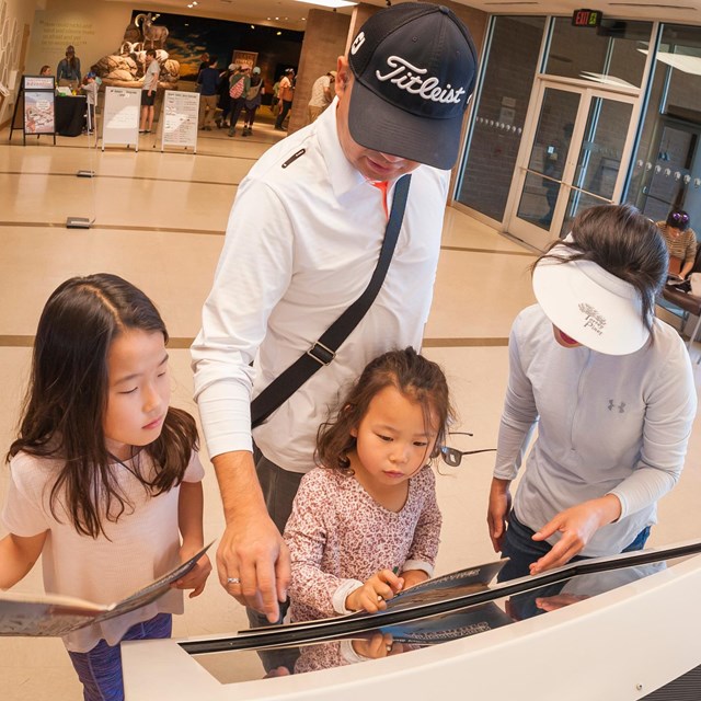 A family looks at a touch screen display with maps in a building with tile floors and glass doors.