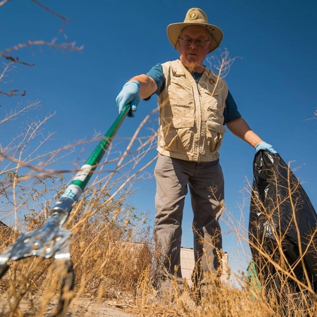A volunteer in tan volunteer uniform reaches towards the camera with a trash picker and black trash.