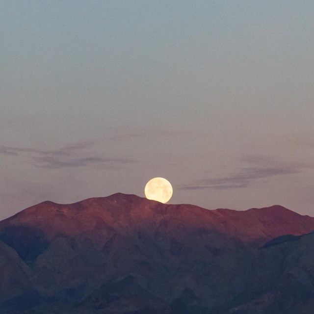 The photograph captures a tranquil twilight scene with a full moon rising just above a mountains.