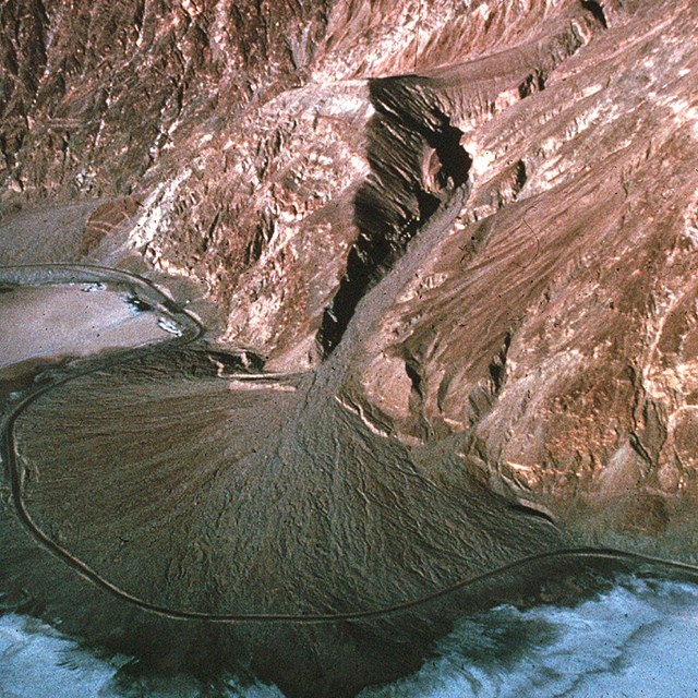 A bird’s eye view of an alluvial fan. The dark brown sediment is in a wide fan shape spilling out.