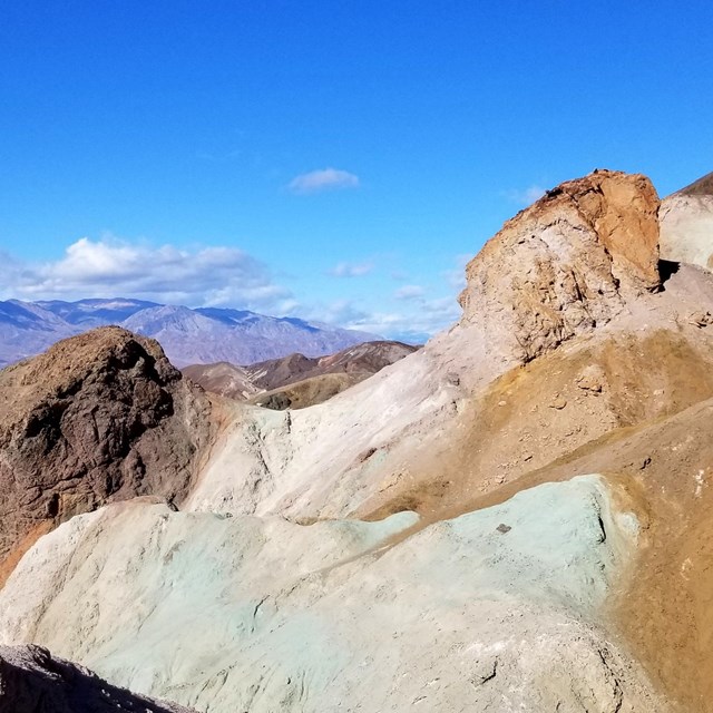 Under a blue sky, a mountainside displays outcrops of volcanic ash deposits in colors.
