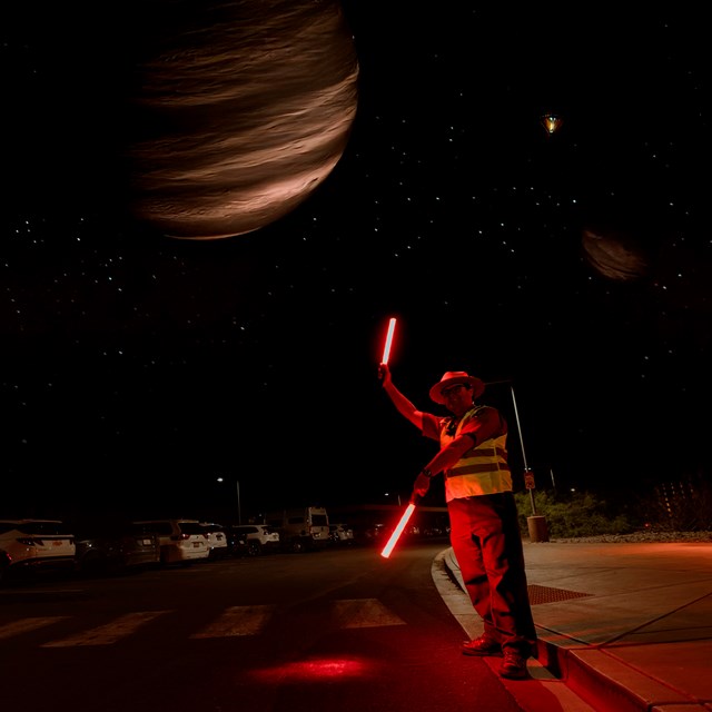 A parking attendant in a reflective vest stands on a street at night, using glowing red batons.