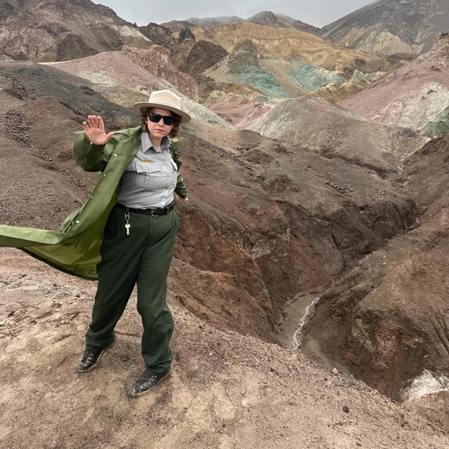 A ranger in a flowing trench coat and sunglasses stands in front of colorful clay hills.