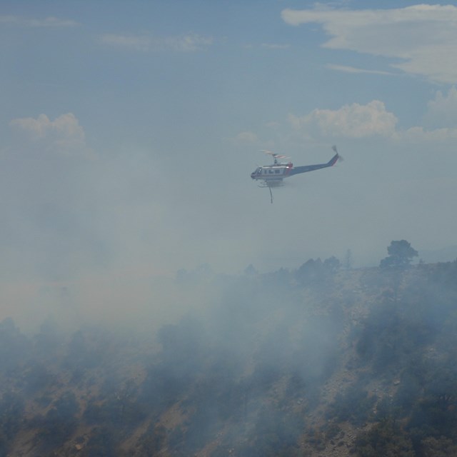 A helicopter flies over a smokey landscape with blue skies above. NPS