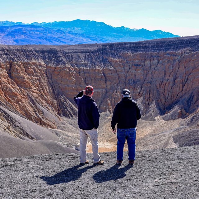 Two visitors gaze out over a large crater, the crater has multiple layers and different colored rock