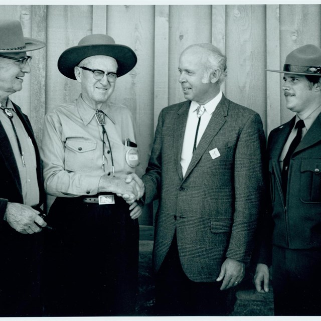 Black and white photo of four men standing in front of wall. Men in the middle are shaking hands.