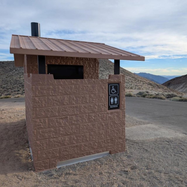 The photograph displays a small, single-unit public restroom made of reddish-brown bricklike materia