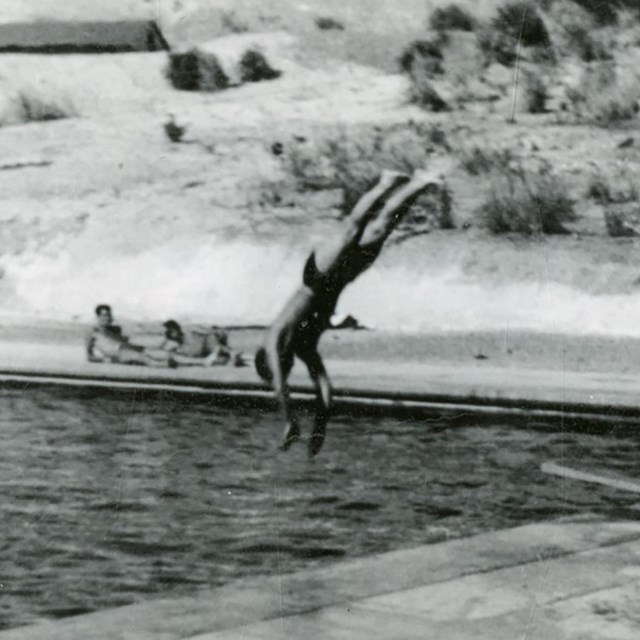 A black an white photo of an individual diving into a pool with others sitting around the edge.