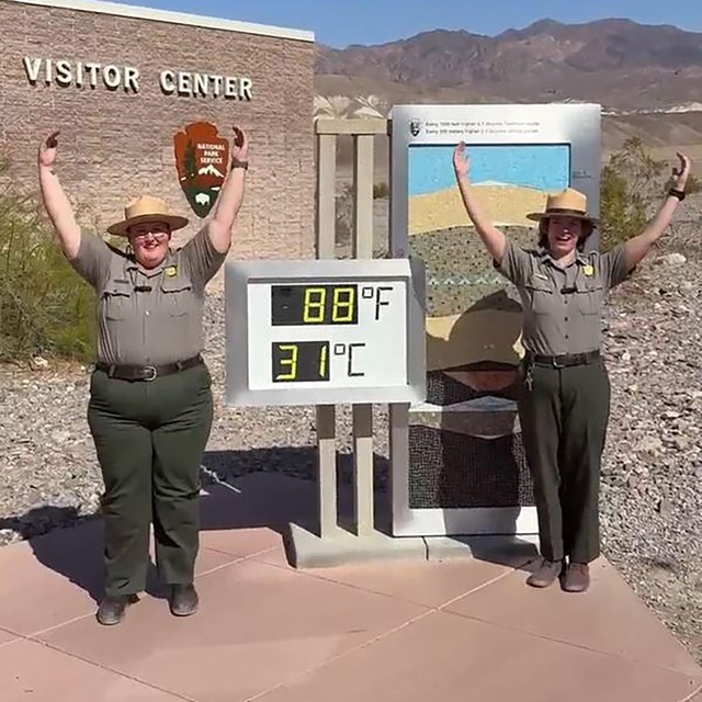 Two park rangers stand outdoors in front of a visitor center and a temperature display.