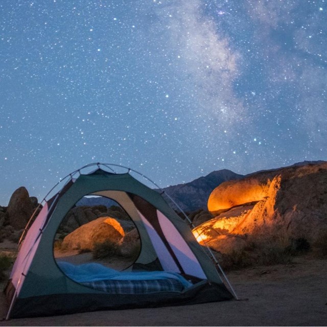 A tent set up under a starry sky among brown, rounded boulders with snowy mountains in the distance.