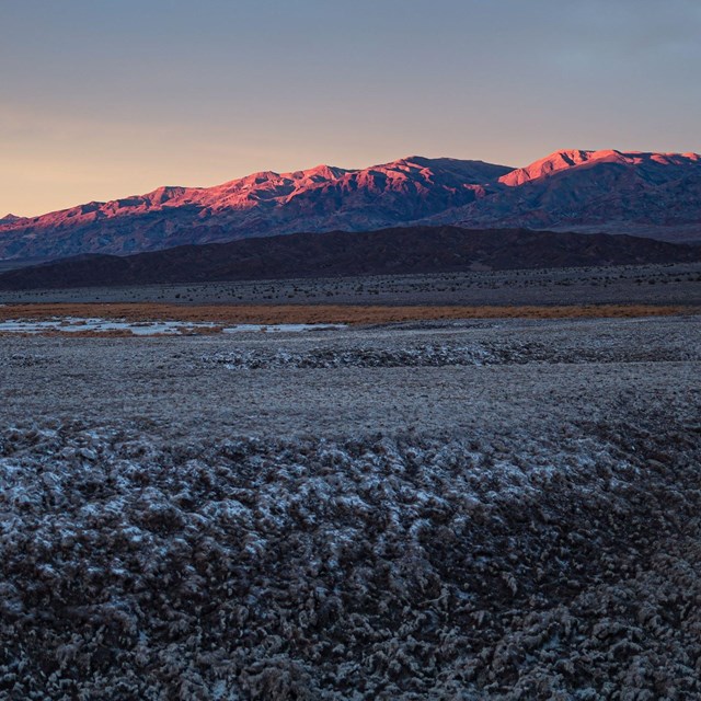 Desert peaks glow pink from sunset, with textured white salt flats in foreground. 