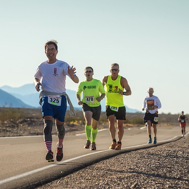  Individuals run along a paved road in a desert landscape with mountains in the background.