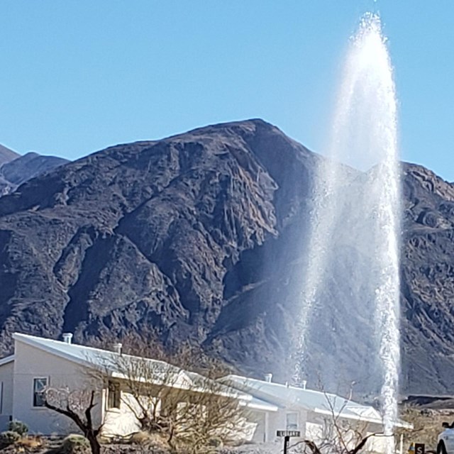 The image shows a sudden and powerful stream of water shooting skyward in front of a single-story ho
