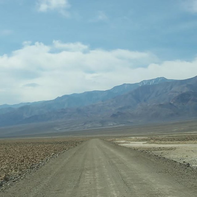 Dirt road in a barren salty landscape with desert mountains and clouds in the distance.