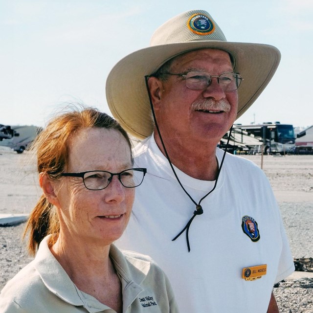 A pair of campground hosts, wearing sandy colored Volunteer uniforms, stand side by side.