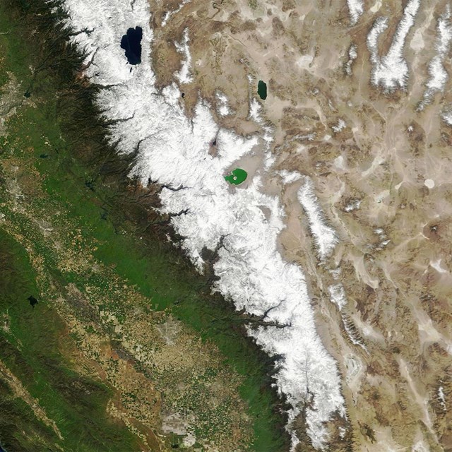 Satellite view of snowy mountains with a lush, green landscape on one side and a brown landscape.