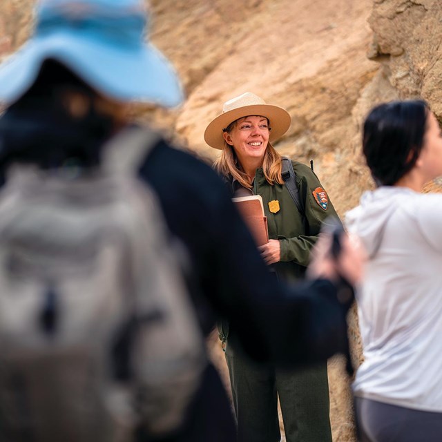 A uniformed ranger speaks to a group of hikers taking pictures, in a yellow/orange canyon with rocky